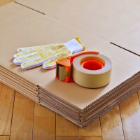 Foto einer Rolle Klebeband, die zusammen mit Arbeitshandschuhen auf ungefalteten Kartons liegt.